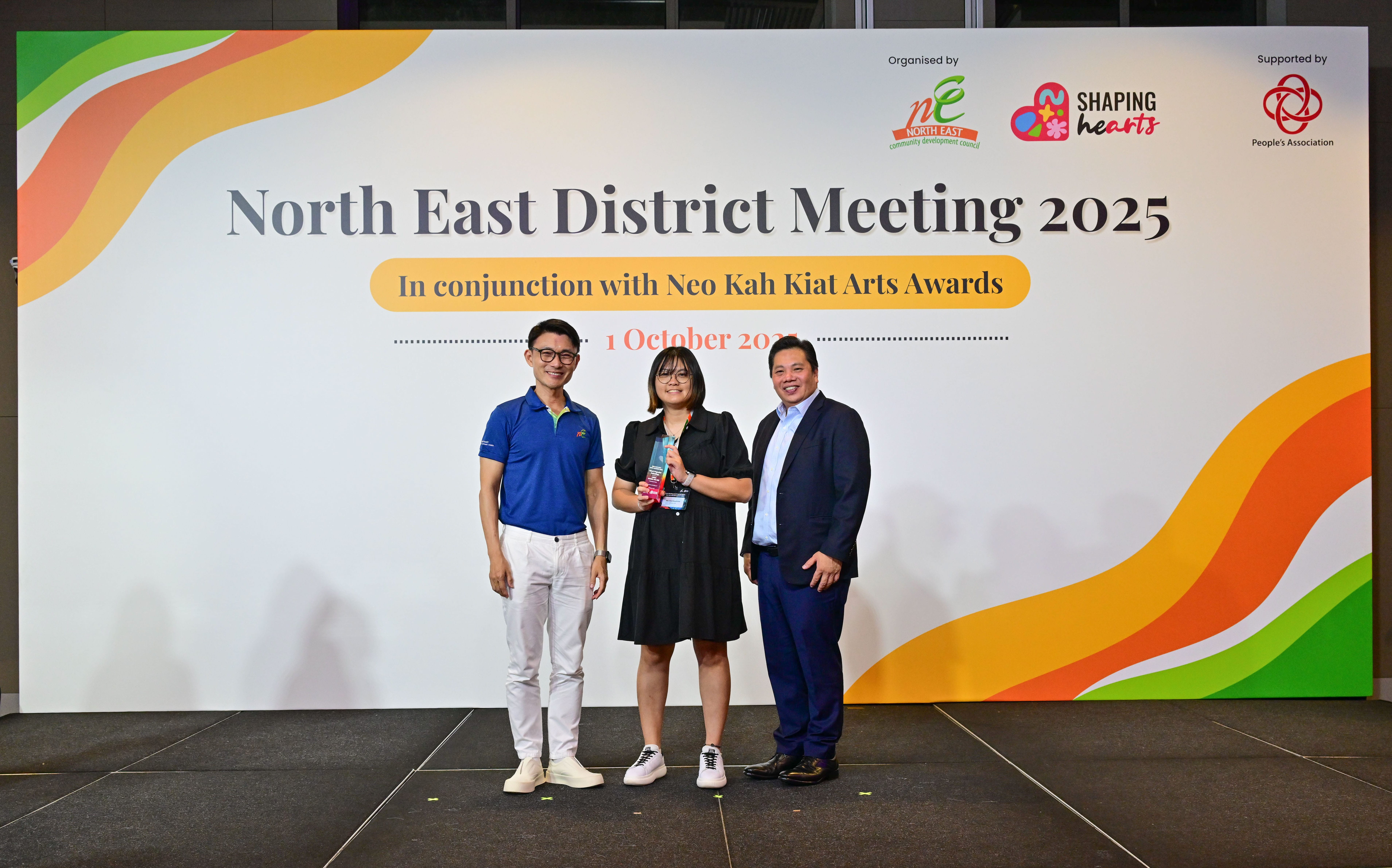 Award recipient standing on stage with the presenter, holding a acrylic plaque and posing for a group photo during the award ceremony, with a backdrop reading ‘North East District Meeting 2025 In conjunction with Neo Kah Kiat Arts Awards'